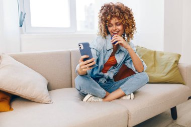 Mutlu genç bir kadın evinde rahat bir koltukta oturuyor, gülümsüyor ve cep telefonunu tutuyor, online oyunlardan ve oturma odasında sohbet etmekten zevk alıyor..
