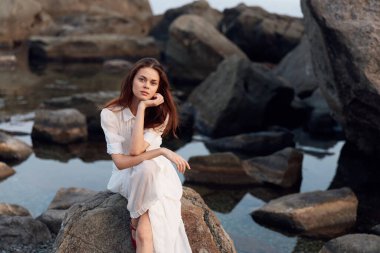 Beyaz elbiseli sakin bir kadın kayalık bir sahilde su kenarında tefekkür ediyor.