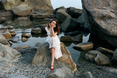 Dalgalı beyaz elbiseli sakin bir kadın okyanus kayalığında oturuyor. Arka planda büyük kayalar var.