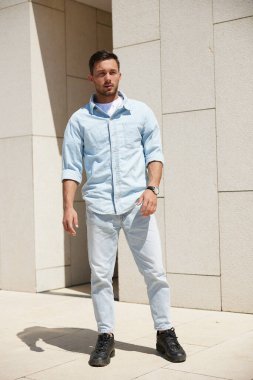 Açık mavi kot gömlek ve kot pantolon giyen özgüvenli bir adam, bej bir binanın arka planında dikiliyor, dümdüz ileriye bakıyor..