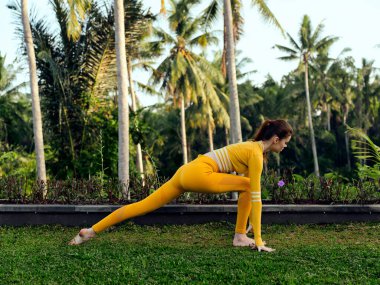 Sarı elbiseli bir kadın arka planda palmiye ağaçları olan yemyeşil çimlerde yoga yapıyor. Huzur ve iç huzuru yayıyor..