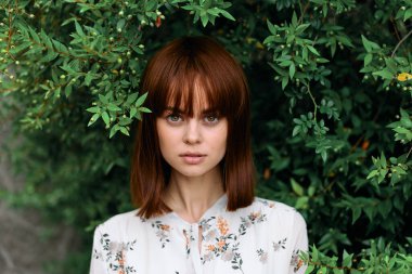 Canlı kızıl saçlı kadın gür yeşil çalıların önünde poz veriyor.