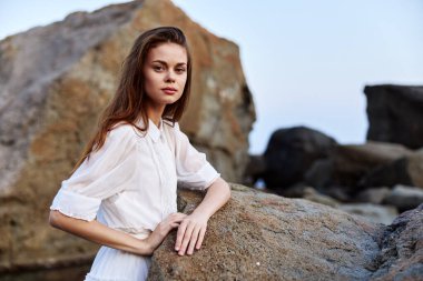 Beyaz elbiseli sakin genç bir kadın deniz kenarındaki kayaya oturmuş, okyanusa bakıyor.
