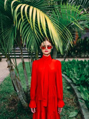 Parlak kırmızı elbiseli ve güneş gözlüklü zarif kadın güneşli bir günde gür palmiye ağaçlarına poz verir..