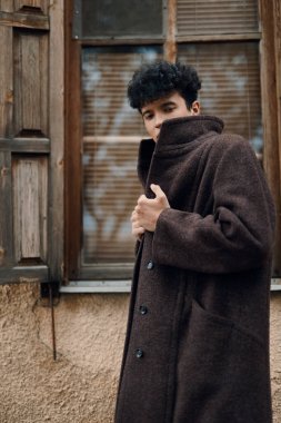 Kafası yana eğilmiş, elleri ceplerinde, kahverengi ceketli, şık bir delikanlı pencerenin önünde duruyor..
