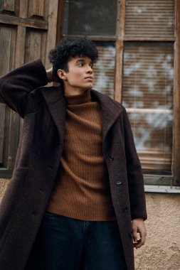 Kahverengi kazaklı ve ceketli manken pencerenin önünde duruyor, elleri başının üstünde, aşağı doğru bakıyor..