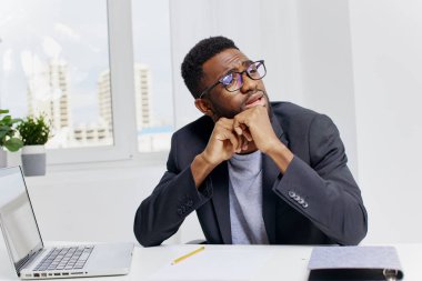 Düşünceli Afro-Amerikan bir adam güncel bir ofiste masasında oturuyor, derin düşüncelere dalmış, dizüstü bilgisayarı ve belgeleriyle..