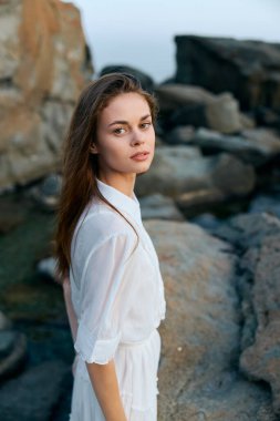 Beyaz elbiseli sakin genç kadın denizin yanındaki kayalık kıyılarda zarifçe poz veriyor.