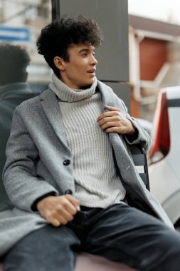 Gri montlu ve beyaz balıkçı yaka kazaklı moda genç adam arabanın önünde bankta oturuyor..