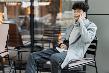 Pencere kenarındaki sandalyede oturan, elinde kahve bardağıyla cep telefonuyla konuşan bir adam. Modern moda yaşam tarzı konsepti.