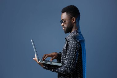 Genç Afro-Amerikan bir adam yoğun bir şekilde bir dizüstü bilgisayar üzerinde çalışıyor, canlı mavi arka planı ile teknoloji ve verimliliği vurguluyor..