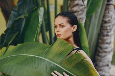 Tropikal bir kadın, cennetteki bir palmiye ağacının önünde büyük yeşil bir yaprak tutuyor.