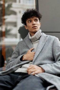 Gri montlu, pencerenin önündeki bankta oturan genç bir adamın moda görüntüsü. Kaplumbağa yakalı süveter ve siyah pantolon giyiyor, kameradan uzağa bakıyor..