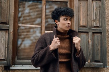 Kahverengi kazaklı ve ceketli moda kadın pencerenin önünde duruyor, elinde ahşap panjurlar ve kapı var..