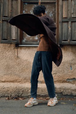 Kahverengi gömlekli, mavi pantolonlu, kolları açık, kafasının üzerinde kahverengi battaniyeyle binanın önünde duran moda adamı..