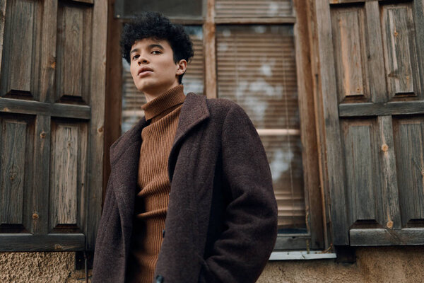 Fashion photo of a stylish young man in a brown coat and turtleneck sweater standing in front of an old window with wooden shutters.