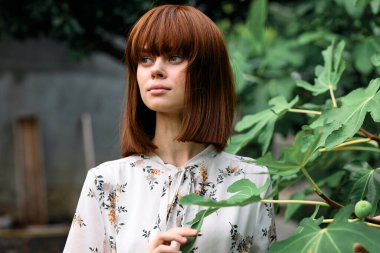 Canlı kızıl saçlı kadın yapraklarıyla yemyeşil ağacın önünde zarif bir şekilde duruyor.