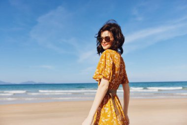 Sarı elbiseli yalnız bir kadın kumlu sahilde sırtı kameraya dönük deniz dalgalarını seyrediyor.