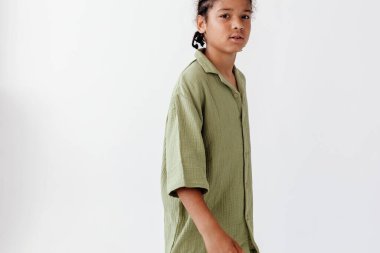 Yeşil gömlekli, siyah pantolonlu, kendine güvenen genç çocuk beyaz duvara karşı poz veriyor..