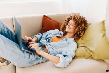 Modern teknoloji ve konforlu oturma odası çevresiyle çevrili rahat bir ofiste dizüstü bilgisayarda çalışan gülümseyen bir kadın..