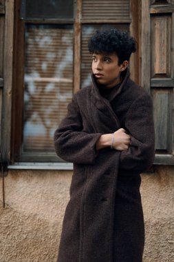 Kahverengi ceketli, elleri kalçasında, pencerenin önünde poz veren ve doğrudan kameraya bakan şık bir genç..