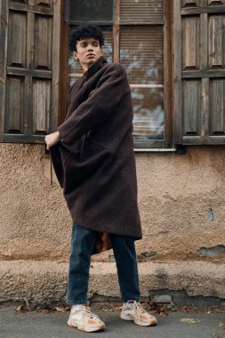 Kahverengi ceketli bir gencin moda fotoğrafı pencerenin önünde duruyor, elleri kalçasında, spor ayakkabı giyiyor..