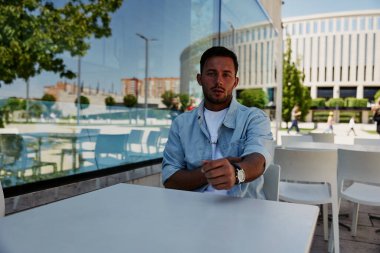 Günlük kıyafetli genç bir adam açık bir kafe masasında oturuyor, kol saatine bakıyor, arkasında modern bir bina ve ağaçlar var..