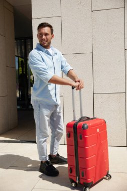 Açık mavi kıyafetli mutlu bir adam kırmızı bavullu, modern binanın dışında duruyor, gülümsüyor ve yan tarafa bakıyor..