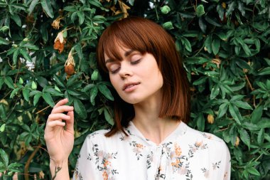 Gözleri kapalı sakin bir kadın, çalılara yaslanırken elinde bir çiçek tutuyor.