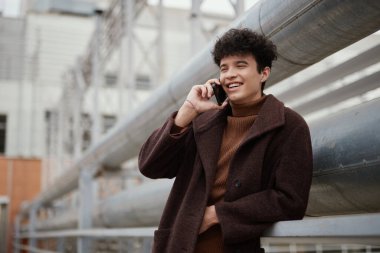 Şehirdeki sanayi binasının üzerinde dururken kahverengi ceketli genç adam cep telefonuyla konuşuyor..