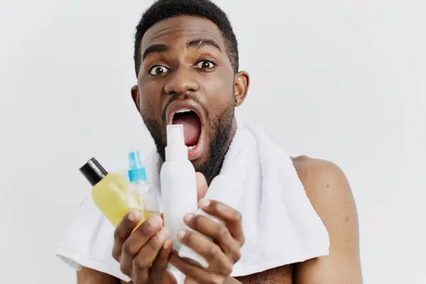 stock image Man with towel draped over head holds shampoo and conditioner bottles, prepared for his daily hair care routine.