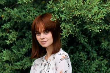 Canlı kızıl saçlı kadın gür yeşil çalıların önünde yapraklarla poz veriyor.