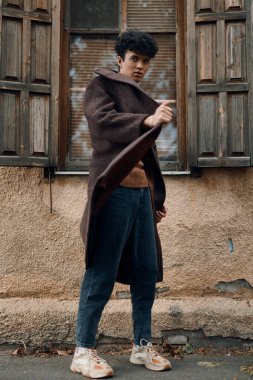 Kahverengi ceketli, mavi kotlu, beyaz spor ayakkabılı, turuncu gömlekli ve kahverengi şapkalı bir adam bir binanın önünde duruyor..