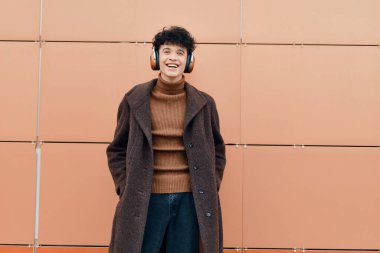 Kahverengi paltolu bir gencin moda portresi, turuncu bir duvarın önünde kulaklıkla duruyor. Yüzünde bir gülümseme var ve elleri cebinde..