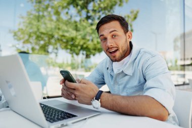 Akıllı telefon tutan mutlu genç bir adam, açık bir kafe masasında dizüstü bilgisayarla oturuyor, heyecan ve şaşkınlığı ifade ediyor. Günışığı, arka planda yeşillik olan modern ortam.