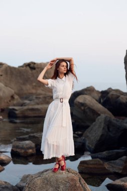 Beyaz elbiseli, kırmızı ayakkabılı, okyanus kıyısında kayalıklarda duran sakin bir kadın.