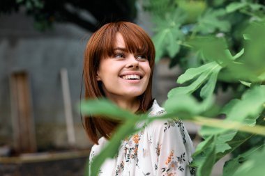 Kızıl saçlı mutlu kadın yemyeşil bitkinin önünde duruyor.