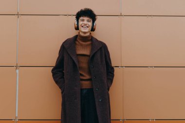 Kulaklık takan, kahverengi ceketli, yüzünde bir gülümsemeyle turuncu bir duvarın önünde duran şık, genç bir adam..