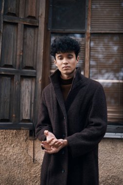 Moda: kahverengi ceketli genç bir adam elleri ceplerinde pencerenin önünde duruyor ve kameraya bakıyor..