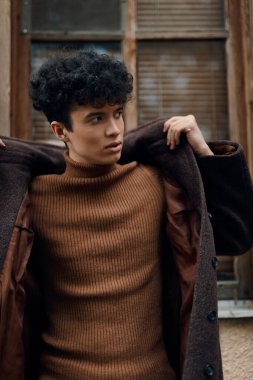 Eski bir şehir binasının önünde kahverengi kazak ve ceket giyen kıvırcık saçlı bir gencin moda fotoğrafı..