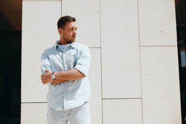 Sıradan bir kot pantolon ve gömlek giyen genç adam, kollarını kavuşturmuş, modern beyaz bir duvarın önünde düşünceli bir şekilde yan tarafa bakıyor..