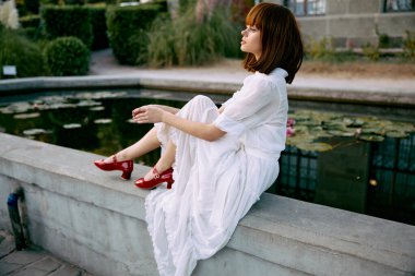 Beyaz elbiseli, kırmızı ayakkabılı, gölün kenarında oturan sakin bir kadın.