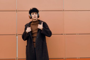 Kulaklık ve palto giyen, elleri kalçasında turuncu duvarın önünde gülümseyen moda adamı..