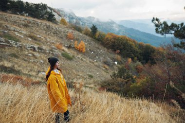 Sarı yağmurluklu bir kadın bulutlu gökyüzünün altındaki görkemli bir dağın önünde çimenli bir tepede duruyor.