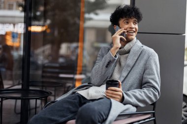 Elinde kahve fincanı olan bir adam arka planda kahve dükkanı olan pencerenin önünde cep telefonuyla konuşuyor. Modern yaşam tarzının moda sahnesi.