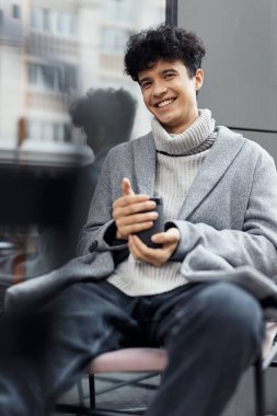 Pencerenin önünde bir fincan kahveyle sandalyede oturan ve kameraya gülümseyen genç bir adamın moda görüntüsü..