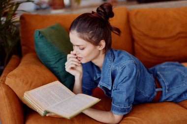 Kitap okuyan kadın, kanepede yüzüstü uzanmış, elleri rahat bir şekilde oturma odasında.