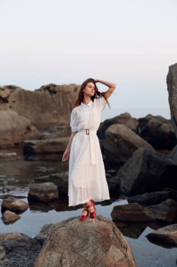 Dalgalı beyaz elbiseli sakin kadın kayalık sahilde poz veriyor, elleri başının üstünde okyanus dalgalarına bakıyor.