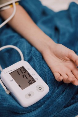Bir kolunda tansiyon monitörü, diğer kolunda kan basıncı kelepçesi olan bir kişi yatakta yatıyor..