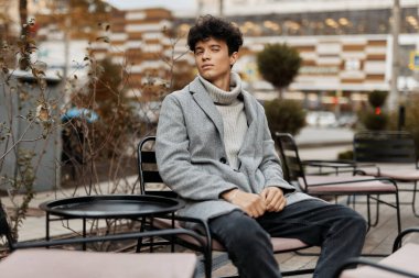 Fashionable young man in a grey coat seated outdoors next to a pink chair. clipart